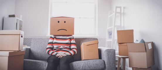 Tired sad woman with cardboard box on her head sitting on the sofa in her new house.