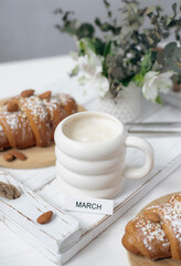 A cup of aromatic cappuccino and a croissant on a white tray. Inscription March