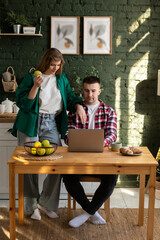 Young couple in the kitchen. A man works at a laptop while his wife leans on his shoulder and watches the process, holding an apple in her other hand.