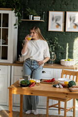 A young woman stands in the kitchen and drinks juice from a glass, looking in front of her. Full height.