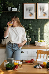 A young woman stands in the kitchen and drinks juice from a glass, looking in front of her. © dmshpak