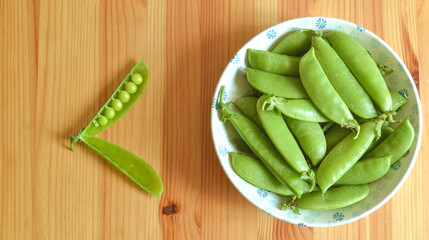 Top view of organic snap peas in an white and blue porcelain bowl with a open pea on the side on a wooden table.