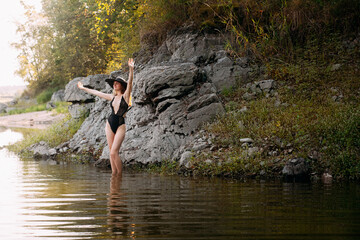 Fototapeta premium A young girl with long blond hair and a beautiful figure, in black openwork lingerie and a black wide hat, stands against the backdrop of a rock, on the banks of a picturesque river.