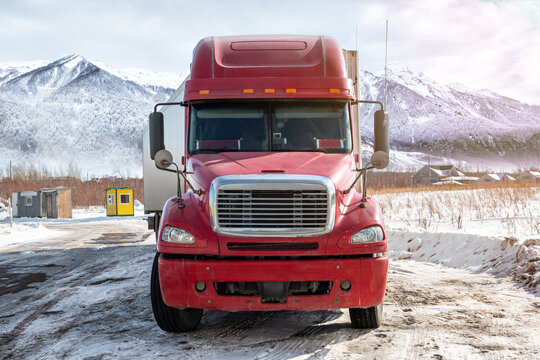 Front view of the red long-distance bonnet truck with a white semitrailer at winter on the background of high scenic mountains