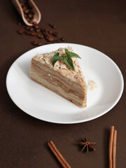 Delicious Napoleon Cake with cream on a white plate decorated with a sprig of mint on a Brown Background. A cinnamon stick, badyan, coffee beans on a brown background. Copy space