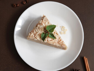 Top view of Delicious Napoleon Cake with cream on a white plate decorated with a sprig of mint on a Brown Background. A cinnamon stick, badyan, coffee beans on a brown background. Copy space