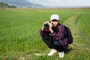 Asian boy wearing plaid shirt and a cap holding a binoculars and looking at the flock of birds feeding on the paddy field and flying in the sky in the paddy field.