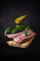 a flying sandwich made from a slice of bread, pork belly, arugula leaves and canned cucumber on a dark gray background with a vignette. fast food concept