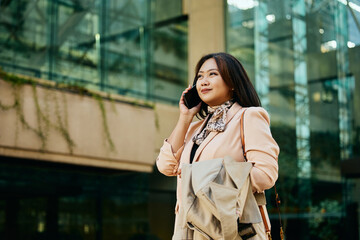 Fototapeta premium Smiling Asian businesswoman communicating on cell phone while walking on street.