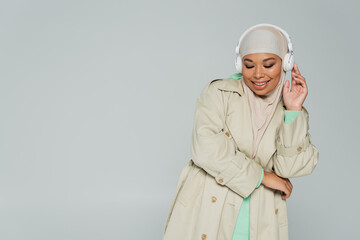 overjoyed multiracial woman in trench coat and hijab listening music in wireless headphones isolated on grey.