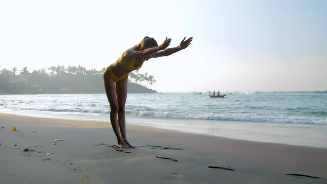 Woman in swimsuit fixes bending pose with stretched hands forward doing yoga by ocean against fishing boat. Lady on wet sand in morning