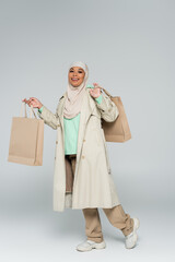 full length of muslim multiracial woman in trench coat and hijab standing with shopping bags on grey background.