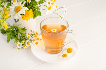 Chamomile tea in a transparent cup on a white wooden background with a bouquet of daisies