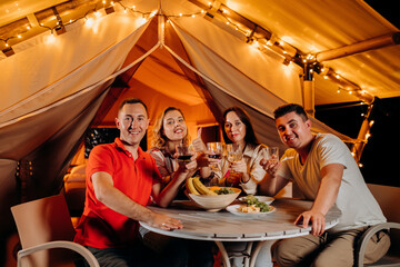 Happy group of friends relaxing in glamping and drinking wine on summer evening near cozy bonfire. Luxury camping tent for outdoor recreation and recreation. Lifestyle concept