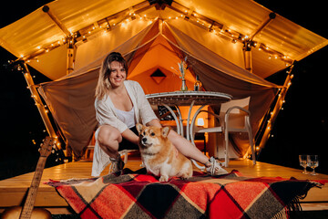 Obraz na płótnie Canvas Happy young woman with her Welsh Corgi Pembroke dog relaxing in glamping on summer evening near cozy bonfire. Luxury camping tent for outdoor recreation and recreation. Lifestyle concept