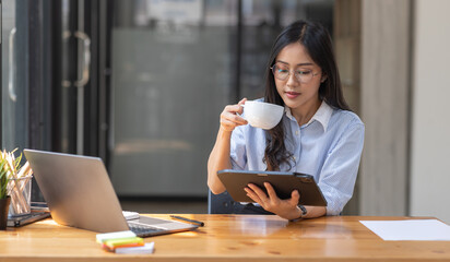 Beautiful young Asian woman working on laptop computer while sitting at the plan office space after working hours, Asian woman drinking coffee