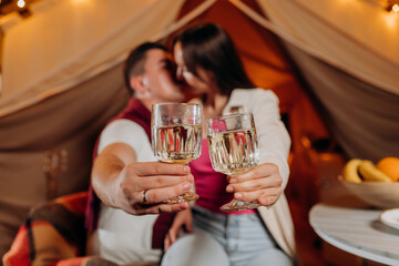 Happy lovely couple relaxing in glamping on summer evening and drinking wine near cozy bonfire. Luxury camping tent for outdoor recreation and recreation. Lifestyle concept