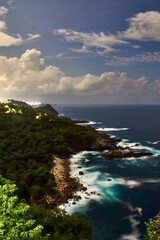 cliff in front of the beach at night with clouds in the sky, and lights of city in the background,...