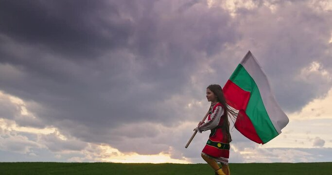 Beautiful bulgarian girl in folklore ethnic costume and bulgarian flag running outdoor on green agriculture field. Freedom, beauty and bulgarian independence and pride concept, 4k video
