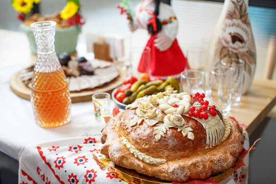 A Ukrainian loaf decorated with rose flowers, a towel, ears of wheat and a bunch of viburnum lies on a towel on the festive table against the background of pickles, cold cuts and a jug of stewed fruit