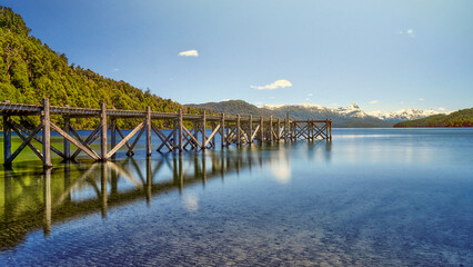 A beautiful pier to the left of the lake in shades of green and blue with a background of mountains...