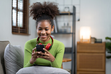  Young smiling African American woman sitting on the sofa at home and texting with her friend on smartphone.
