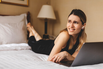 Happy woman using laptop making video call lying on bed at home in bedroom look at side. Modern communication. Freelancer lady browsing internet. Technology and freelance.