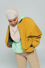 trendy multiracial woman in yellow bomber jacket and hijab posing with hand in pocket isolated on...