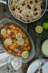 Salmon bites baked in oven with lime and rosemary, pasta in pan and cream sauce in pot on rustic wooden table.