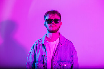Handsome stylish man with trendy sunglasses in a fashion denim jacket on a pink background with creative colorful pink and blue light