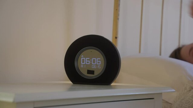 Female turning off the ringing digitall alarm clock on a bedside table at 6 in the morning