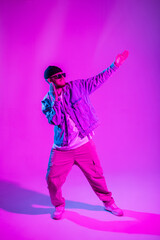 Fashionable handsome dancer hip hop guy with trendy sunglasses and cap in stylish denim clothes dancing in studio with creative pink and blue light