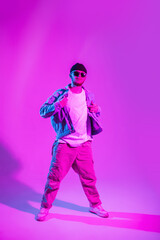 Stylish handsome young male dancer with fashion sunglasses and hat in trendy denim clothes is dancing in colored studio with pink and blue light