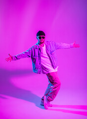 Fototapeta na wymiar Fashionable hipster dancer man with hat and sunglasses in fashion stylish denim clothes with jeans jacket dance in creative colorful studio with pink and blue neon light