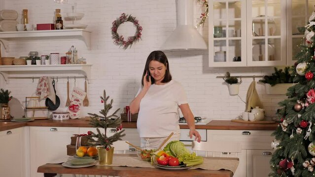Happy pregnant woman talking on the phone in the kitchen on christmas eve. Nearby is a decorated Christmas tree.
