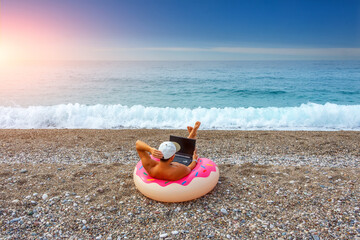 Guy in a cap sits in an inflated circle in the form of a donut with pink icing on the beach and works in a laptop online. The concept of employment on vacation, combining work and leisure.