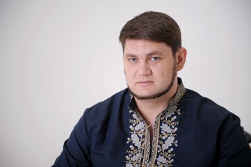 Obraz premium stern look of blue eyes handsome man in Ukrainian embroidered shirt looks straight into frame he has light beard compressed lips thick hair a blue shirt and embroidery on white background close-up