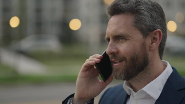 Businessman dressed in formal suit outside using mobile phone. Middle aged man walking on street and speaking phone and smiling. Mature man talking on phone. Businessman in formal suit using phone.