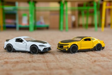 Two toy models of sports cars stand on piled brown carpet against abstract blurred background.
