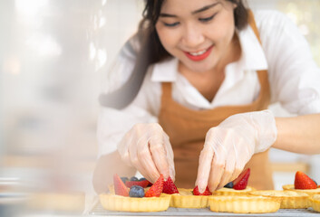 Obraz na płótnie Canvas Young beautiful woman is baking in kitchen , bakery and coffee shop business. Hands in cooking gloves decorating freshly baked tart with blueberries strawberry fresh fruit. homemade bakery at home.