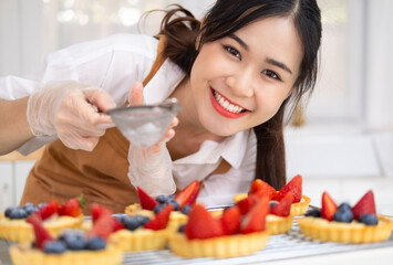 Icing sugar sprinkled with colander. Woman making fruit tart ,sprinkling powdered sugar on top. Smiling House wife wearing apron hands in cooking gloves making finishing touches on birthday dessert.
