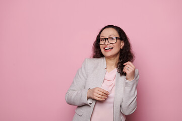 Happy multi-ethnic emotional middle-aged woman, wearing black trendy eyeglasses and elegant light gray suit, laughing looking at camera on isolated pink background. People< ophthalmology and fashion