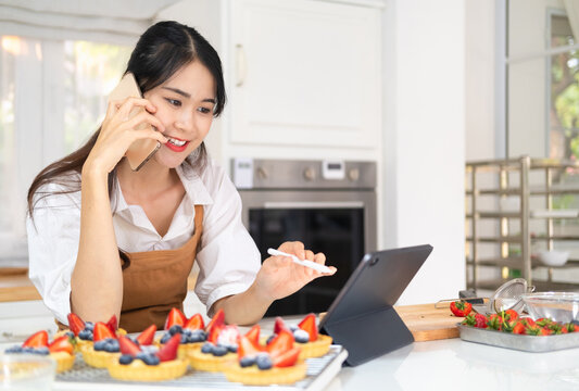 Baker working in home bakery shop using digital tablet computer and talking on cell phone with customer and writing down order details. Owner bakery working pointing order delivery in digital tablet.