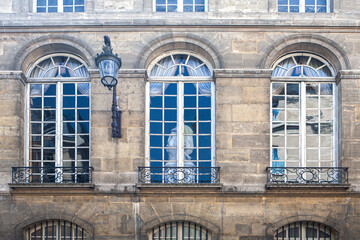 Fototapeta na wymiar Paris ancient stone building facade with three rows of French windows, stucco fretwork, small wrought iron balconies and wall lamp.