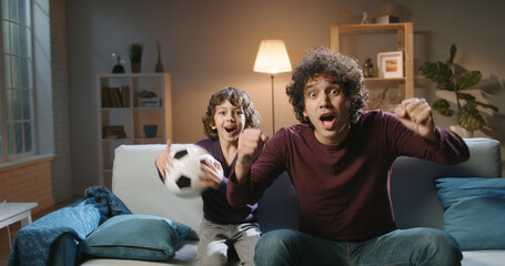 Authentic young cute asian brothers with curly hair watching soccer together, father and son...