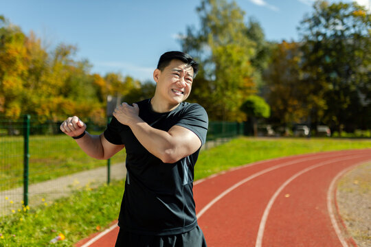 Asian sportsman stretches his shoulder, man in the stadium after running and active physical exercises stretches his arm joints, has shoulder pain, arm muscle spasm.