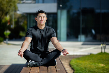Asian man meditates in the lotus position, man in the park near the lake performs yoga exercises, sportsman rests with a closed chin after active physical exercises and running.