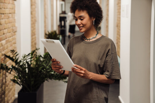 African woman holding clipboard and reading document while standing in coworking - Powered by Adobe