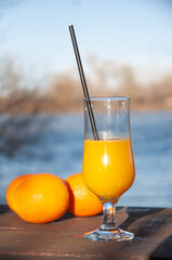 A glass of orange juice with a straw and orange fruit on a wooden table in nature. Behind is water and forest.