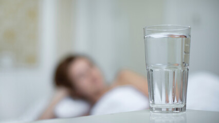 Young woman in the bed in bedroom at home in the morning lying under white blanket and drinking pure fresh water from glass 